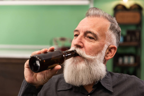 Anciano caucásico barbudo, modelo barbudo, en un bar retro hipster bebe una botella de cerveza, foto retrato. - Foto, Imagen