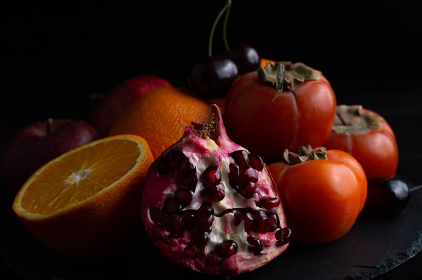 Still life of half a juicy pomegranate, persimmon, apples, orange and sweet cherry on a black background. - Photo, Image