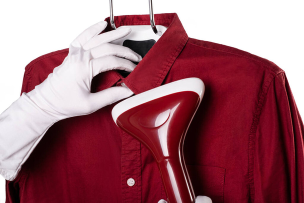 un hombre planchando una camisa roja con un vapor. manos en guantes blancos sostienen un vapor para la ropa y en casa. limpieza y lavado - Foto, Imagen