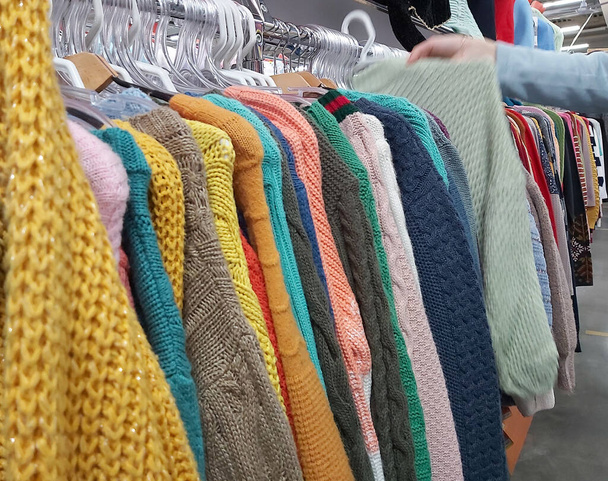 Девушка выбирает теплые трикотажные свитера на вешалках в магазине. - Фото, изображение
