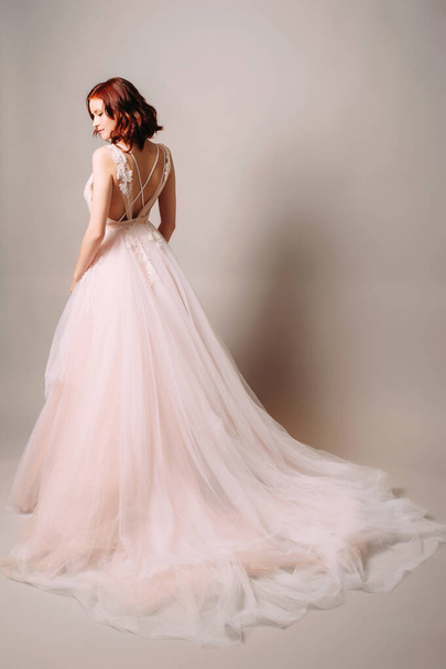 トレンディなウェディングドレス。スタジオに立つ長い列車とピンクのチュールのウェディングドレスのジンジャーブライド。グレーの背景にバック詳細と紫のウェディングドレスの美しい花嫁。ウェディングファッション｜Rear view - 写真・画像