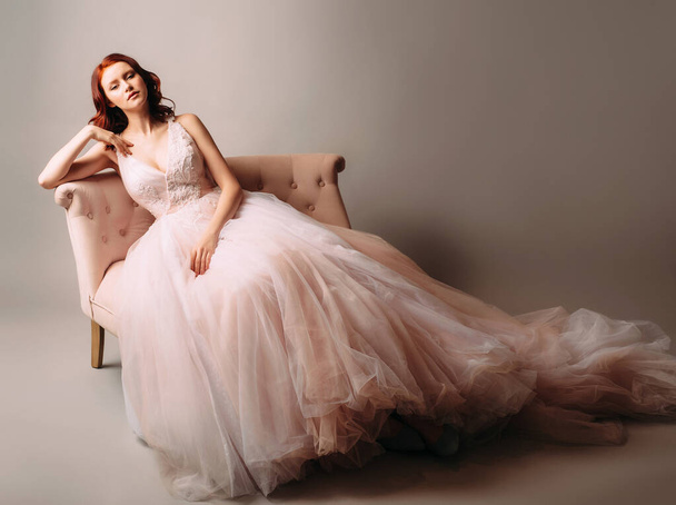 Красива наречена в рожевій весільній сукні, що сидить у вінтажному дивані. Студійний портрет на нейтральному фоні
.  - Фото, зображення