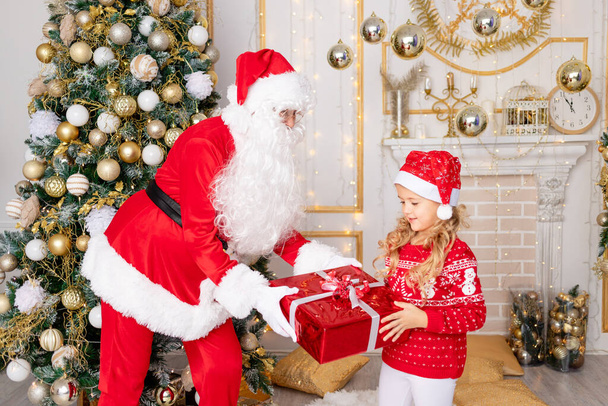 Santa Claus gives a baby girl a gift at the Christmas tree, the concept of new year and Christmas - Photo, Image