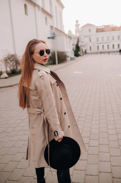 Stylish young woman in a beige coat and a black hat in his hands and glasses on a city street. Women's street fashion. Autumn clothing.Urban style - Fotoğraf, Görsel