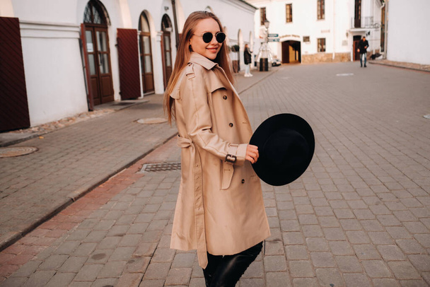 Stylish young woman in a beige coat and a black hat in his hands and glasses on a city street. Women's street fashion. Autumn clothing.Urban style - Foto, imagen