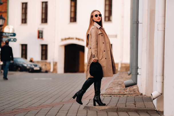Stylish young woman in a beige coat and a black hat in his hands and glasses on a city street. Women's street fashion. Autumn clothing.Urban style - Zdjęcie, obraz
