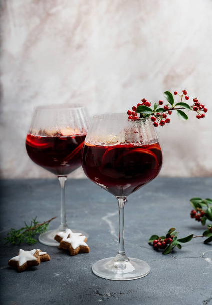 Σπιτικό ζεστό κρασί σερβιρισμένο σε ποτήρια κρασιού διακοσμημένα με χριστουγεννιάτικα μπισκότα, κόκκινα μούρα, πεύκα και ξηρά πορτοκάλια.  - Φωτογραφία, εικόνα