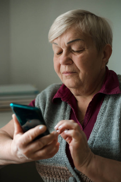 Une vieille femme utilise un téléphone portable. Une femme âgée appelle sur un smartphone alors qu'elle est assise dans une pièce à la maison. - Photo, image