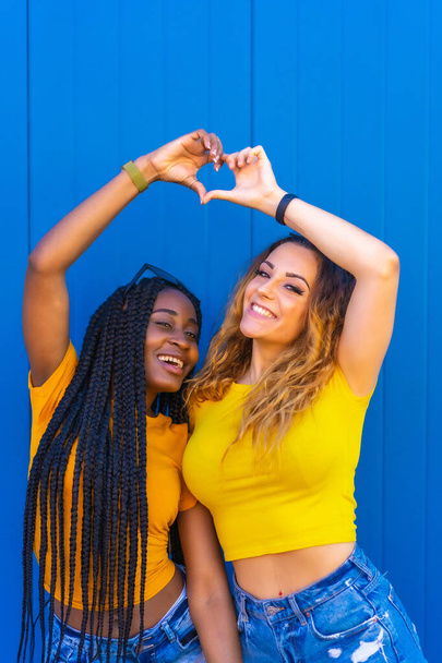 Lifestyle, teenage girl friends reflecting love with each other on a blue wall background dressed in yellow t-shirts. Black girl with long braids and blonde Caucasian girl. - Foto, Imagen