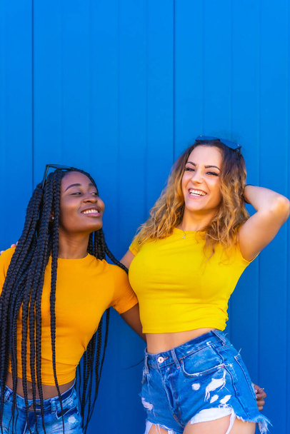 Lifestyle, teenage girl friends reflecting love with each other on a blue wall background dressed in yellow t-shirts. Black girl with long braids and blonde Caucasian girl. - Photo, Image