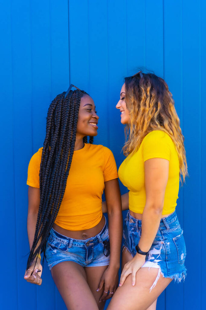 Lifestyle, teenage girl friends reflecting love with each other on a blue wall background dressed in yellow t-shirts. Black girl with long braids and blonde Caucasian girl. - Fotografie, Obrázek