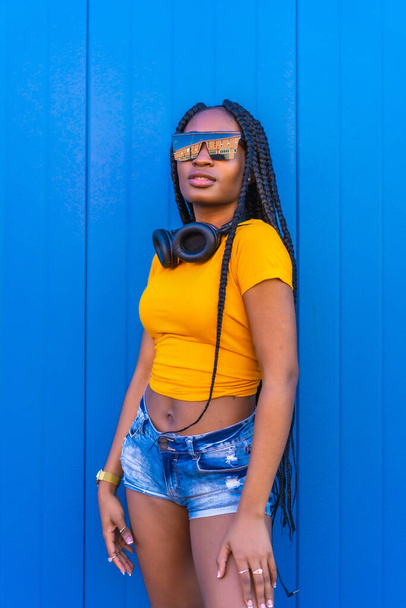 Lifestyle, black girl with long braids, in a yellow shirt and sunglasses on a blue background. Portrait of young dj with headphones smiling while posing for camera, techno, house music - Fotografie, Obrázek