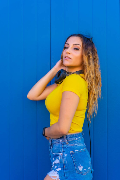 Lifestyle, blonde Caucasian girl with yellow t-shirt on a blue background. Dj girl with headphones promo photo for techno, house party or session poster - Фото, изображение