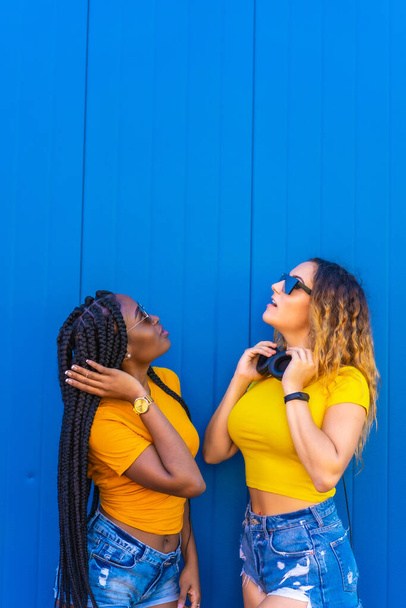 Lifestyle, black girl and blonde Caucasian girl in yellow dresses on a blue background. Looking up, with headphones and sunglasses - Zdjęcie, obraz
