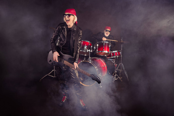 Full size photo of two cool aged lady man rock group performing concert drum instruments play solo guitar fog in air wear trendy rocker leather outfit bandana isolated black color background - Zdjęcie, obraz
