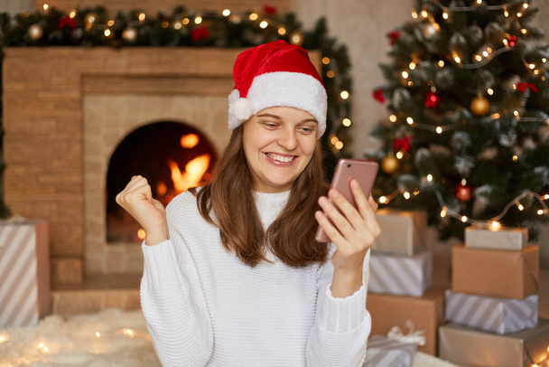 Photo of happy smiling girl reads x-mas news on smartphone, raises fists while sitting in house with christmas decoration and fireplace on background, lady wearing santa hat and sweater. - 写真・画像