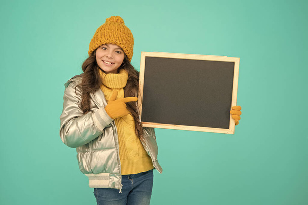 Promotion concept. Kid cheerful promoting. Smiling girl wear winter outfit show blank chalkboard copy space. Informing kids community. Kid with blackboard. Promoting product. Child promoting event - Photo, image