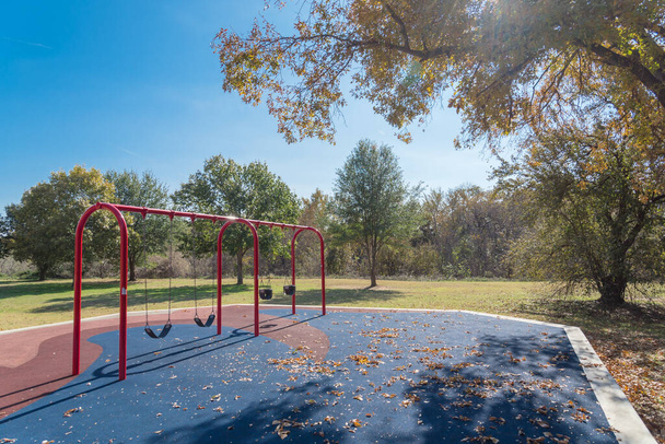 Czerwona huśtawka na publicznym placu zabaw w pobliżu parku przyrody jesienią z kolorowymi liśćmi jesieni w Flower Kopiec, Texas, Ameryka. Upadłe liście suszone na podłożu gumowym. - Zdjęcie, obraz