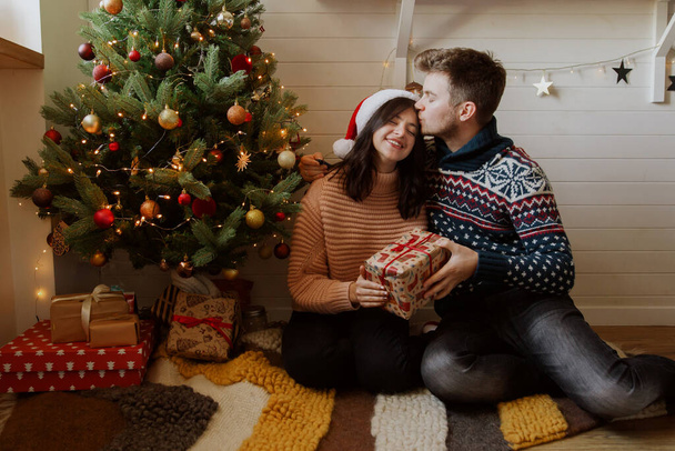 Stylish happy couple exchanging christmas gifts under christmas tree with presents and lights in festive decorated room. Young family kissing and holding present, magic moments. Merry Christmas! - Foto, imagen