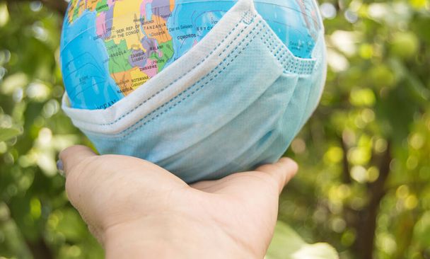 Hand holding a model of the globe in a medical mask, coronavirus control concept, outdoor, summer, blurred background of plants - Photo, Image