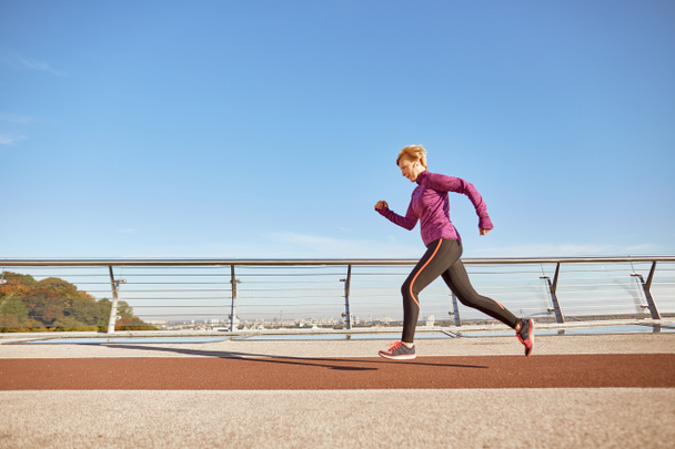 Zdrowie jest najważniejsze. Pełne ujęcie zmotywowanej, aktywnej, dojrzałej kobiety w odzieży sportowej biegającej na świeżym powietrzu w słoneczny dzień. Widok boczny - Zdjęcie, obraz