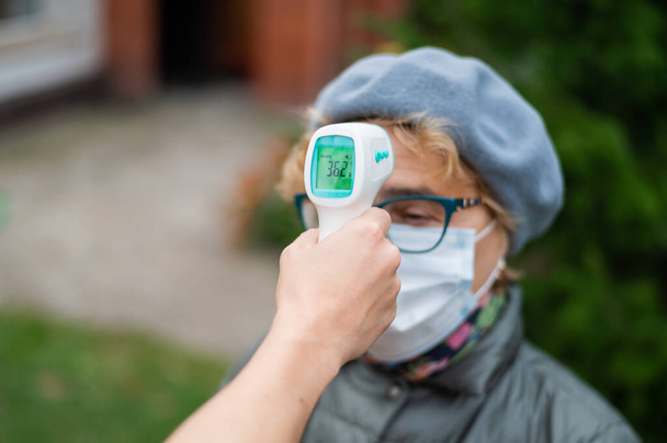 医師は、屋外で非接触赤外線温度計を備えたマスクを着用する高齢女性の温度を測定します - 写真・画像