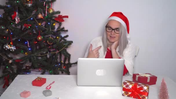 Karácsonyi online család gratulálok. Mosolygó lány otthon használ laptop számítógép notebook videohívás barátok és szülők Mikulás kalap. Egy nő kamerát néz és integet az üdvözlő kezével. 4k videó - Felvétel, videó