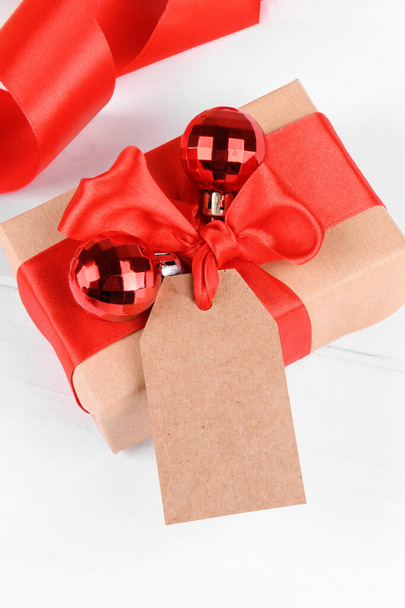 Christmas gift tag mock up with gift box wrapped in craft recycled paper with red ribbon bow on a white backgroud - Photo, Image