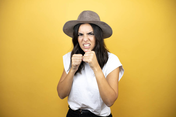 Beautiful woman wearing casual white t-shirt and a hat standing over yellow background Punching fist to fight, aggressive and angry attack, threat and violence - Foto, immagini