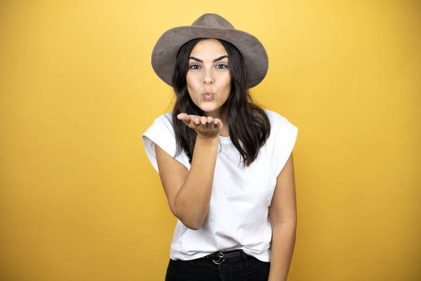 Beautiful woman wearing casual white t-shirt and a hat standing over yellow background looking at the camera blowing a kiss with hand on air being lovely and sexy. Love expression. - Fotografie, Obrázek