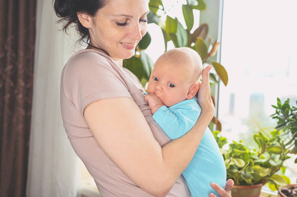 Όμορφη μητέρα που κρατάει αγκαλιά τον μικρό χαριτωμένο νεογέννητο γιο της. Παιδικές εκφράσεις προσώπου. Οικογένεια στο σπίτι δίπλα στο παράθυρο. Υγιές παιδί, έννοια του νοσοκομείου και ευτυχισμένη μητρότητα. Φυτώριο. - Φωτογραφία, εικόνα