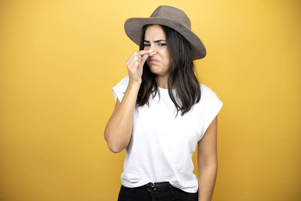 Beautiful woman wearing casual white t-shirt and a hat standing over yellow background smelling something stinky and disgusting, intolerable smell, holding breath with fingers on nose - Foto, Imagem