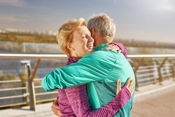 Ευτυχισμένοι μαζί. Ενεργό ώριμο οικογενειακό ζευγάρι στα αθλητικά αγκαλιάζει μετά από προπόνηση στο πάρκο της πόλης σε ένα ηλιόλουστο πρωινό. Χαρούμενο ζευγάρι ηλικιωμένων που στέκονται μαζί στην ύπαιθρο - Φωτογραφία, εικόνα