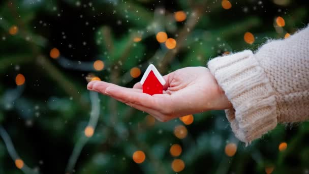 Mujer sosteniendo pequeña casa roja en la mano en las luces de Navidad y abeto en el fondo - Metraje, vídeo
