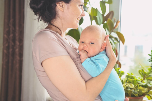 Beautiful mother holding in arms her little cute newborn infant son. Baby facial expressions. Family at home by the window. Healthy child, concept of hospital and happy motherhood. Nursery. - Photo, Image