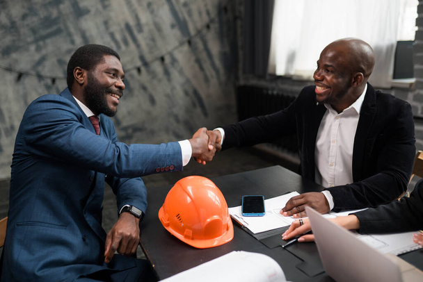 Two African Americans have signed an agreement and are shaking hands. Businessmen are sitting at an office table with an ornamental helmet and documents - Zdjęcie, obraz