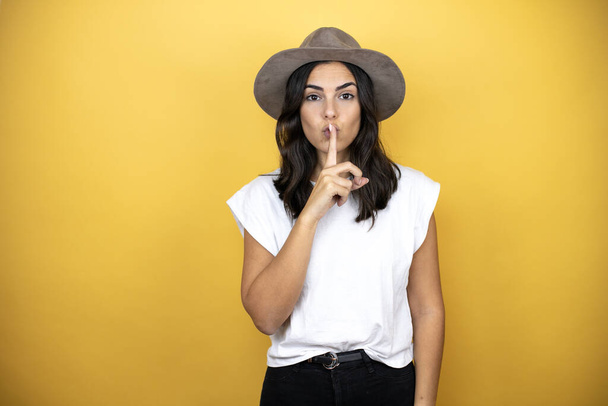 Beautiful woman wearing casual white t-shirt and a hat standing over yellow background asking to be quiet with finger on lips. Silence and secret concept. - Fotografie, Obrázek