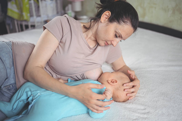 Молодая мать лежит с новорожденным милым младенцем в синем комбинезоне на кровати, обнимает и кормит грудным молоком. Здоровый ребенок, концепция больницы и счастливое материнство. Детская комната. - Фото, изображение