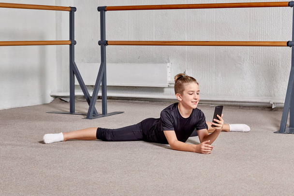 A young man is sitting on the splits in the gym and holding a smartphone. - Фото, изображение