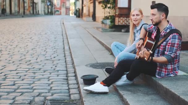 Νεαροί τραγουδιστές του δρόμου στο παλιό τμήμα της πόλης κάθονται στο πεζοδρόμιο - Πλάνα, βίντεο
