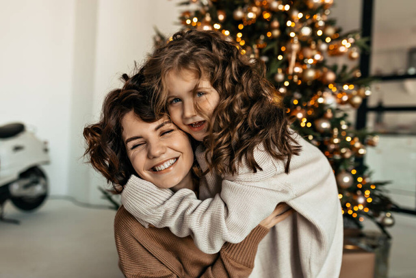 Feliz mujer encantadora con su pequeña hija linda con el pelo ondulado abrazando y favoreciendo la diversión frente al árbol de Navidad. Foto de alta calidad - Foto, Imagen