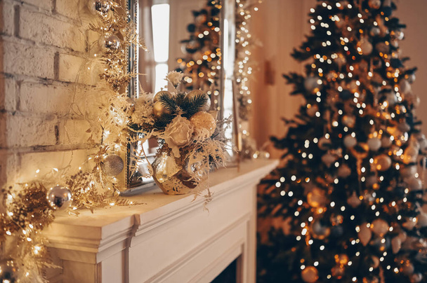 White classic fireplace with New Year decorations against the background of Christmas decorated tree with sparkling garlands. Fragment of New Year festive living room interior at night - Foto, afbeelding