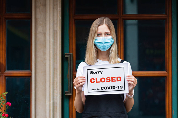 Caucasian waitress woman wearing medical mask holds SORRY WE'RE CLOSED. Coronavirus pandemic. Government shutdown of restaurants, shopping stores, non essential services. High quality photo - Valokuva, kuva