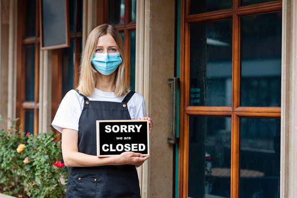Caucasian waitress woman wearing medical mask holds SORRY WERE CLOSED. Coronavirus pandemic. Government shutdown of restaurants, shopping stores, non essential services - Foto, imagen