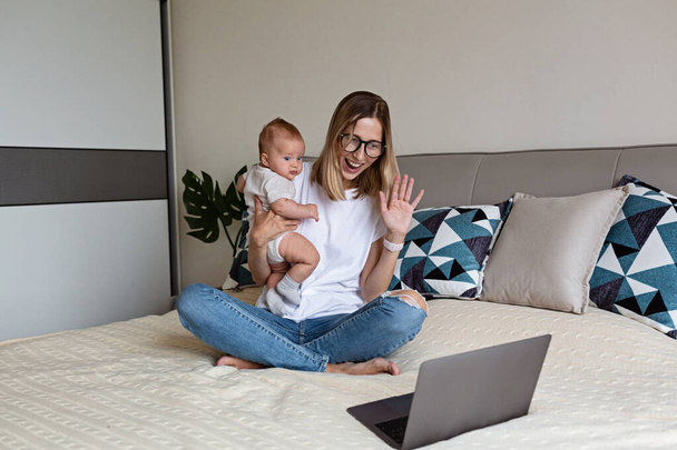 Telecommuting in quarantine. Mother showing baby to friends at the video chat. Stylish young freelancer working from home. Social distancing during coronavirus covid-19 pandemic concept. - Foto, imagen
