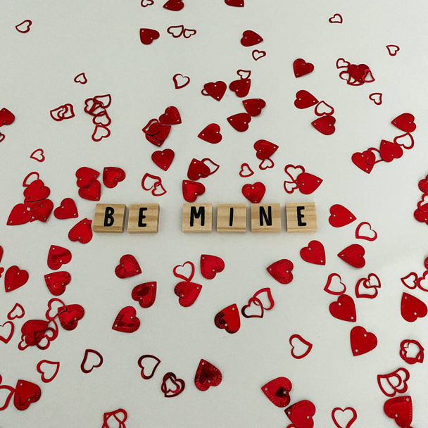 Houten ambachtelijke letters die Be Mine spellen met rode hart confetti op een witte achtergrond. Concept liefde, romantiek, bruiloft, verloving, verjaardag en Valentijnsdag. - Foto, afbeelding