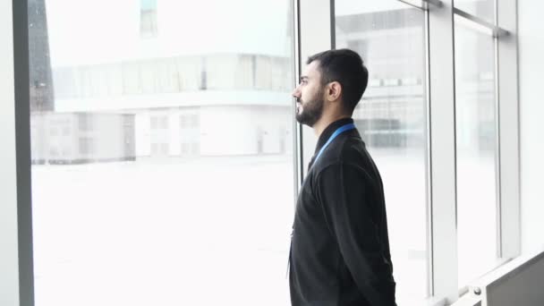 Side view of a pensive young man standing by the window. Media. Businessman with a blue name tag of a forum participant looking at falling snow behind the window. - Footage, Video