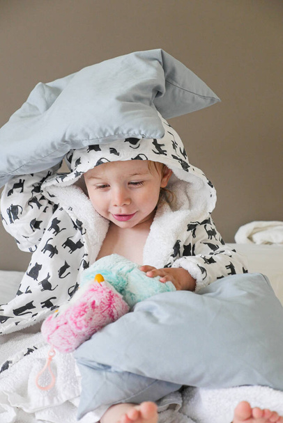 Criança vestindo roupão com capuz brincando com travesseiro na cama. Tiro médio. Vertical.  - Foto, Imagem