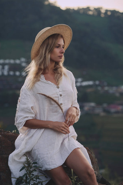 The blonde in a linen dress and straw hat sits on a viewpoint in the mountains - Zdjęcie, obraz