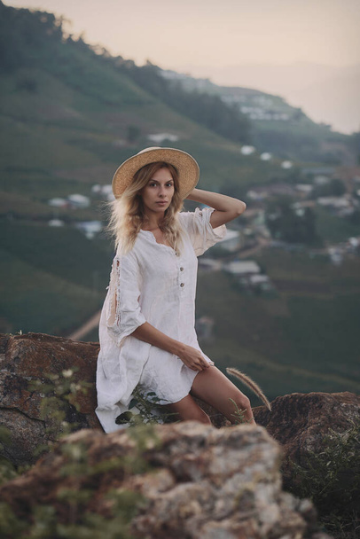 The blonde in a linen dress and straw hat sits on a viewpoint in the mountains - 写真・画像